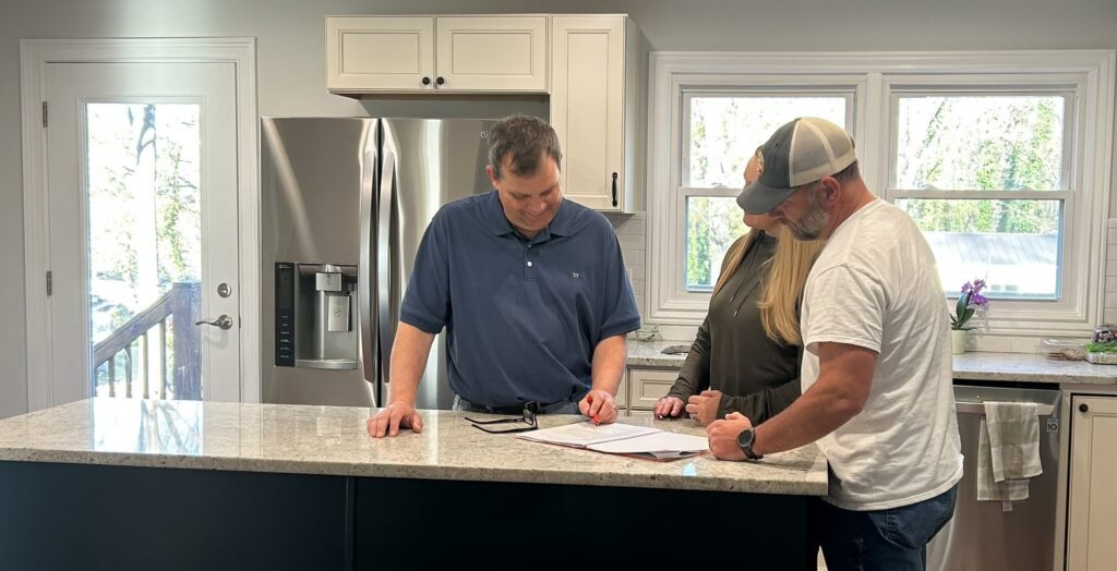 Three people are standing in a kitchen looking at paperwork.